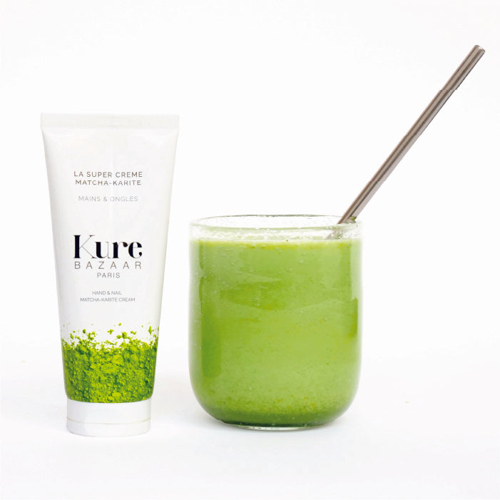A bottle of matcha shea butter nail balm by Kure Bazaar with a glass of matcha tea and a metal straw