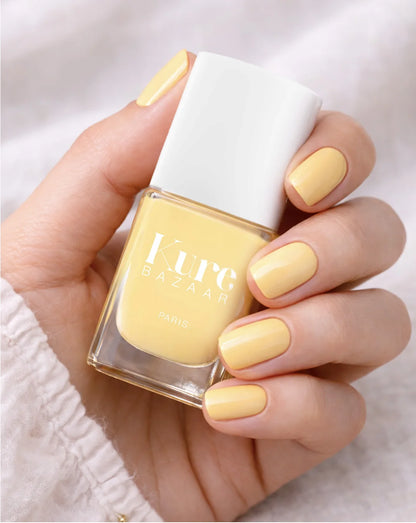 A hand holding a bottle of Kure Bazaar nail polish, showing the pale pastel yellow shade applied to the fingernails against a soft white fabric background.